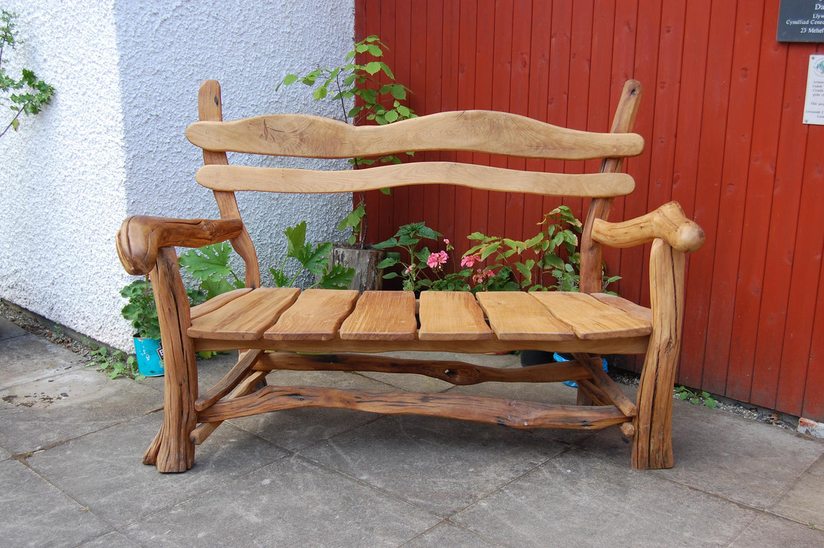 A rustic handcrafted wooden bench made from natural tree trunks and slatted backrest, positioned outdoors against a red wooden fence and a white stucco wall.