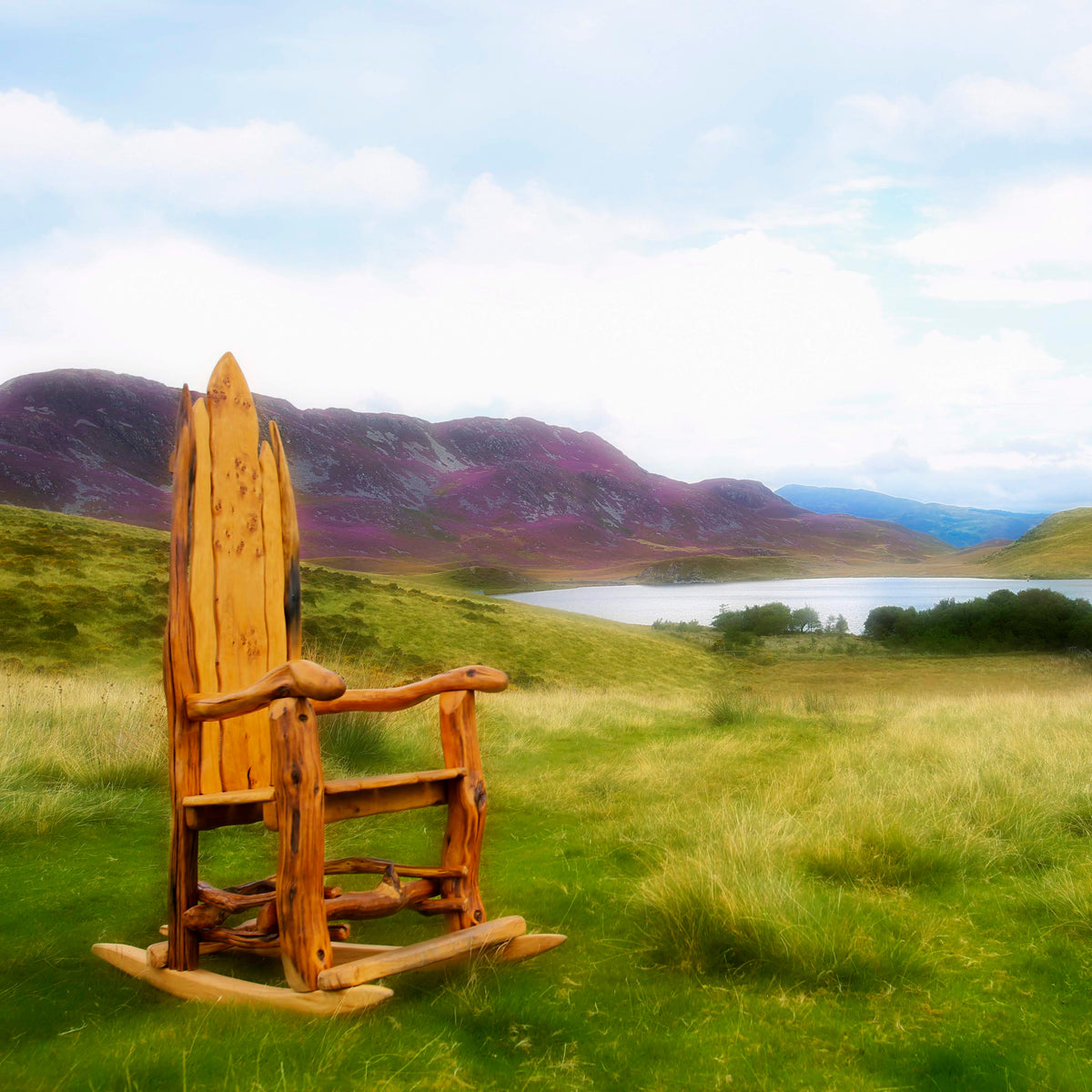 A rustic wooden rocking chair placed in a grassy field with a scenic mountain lake landscape in the background.