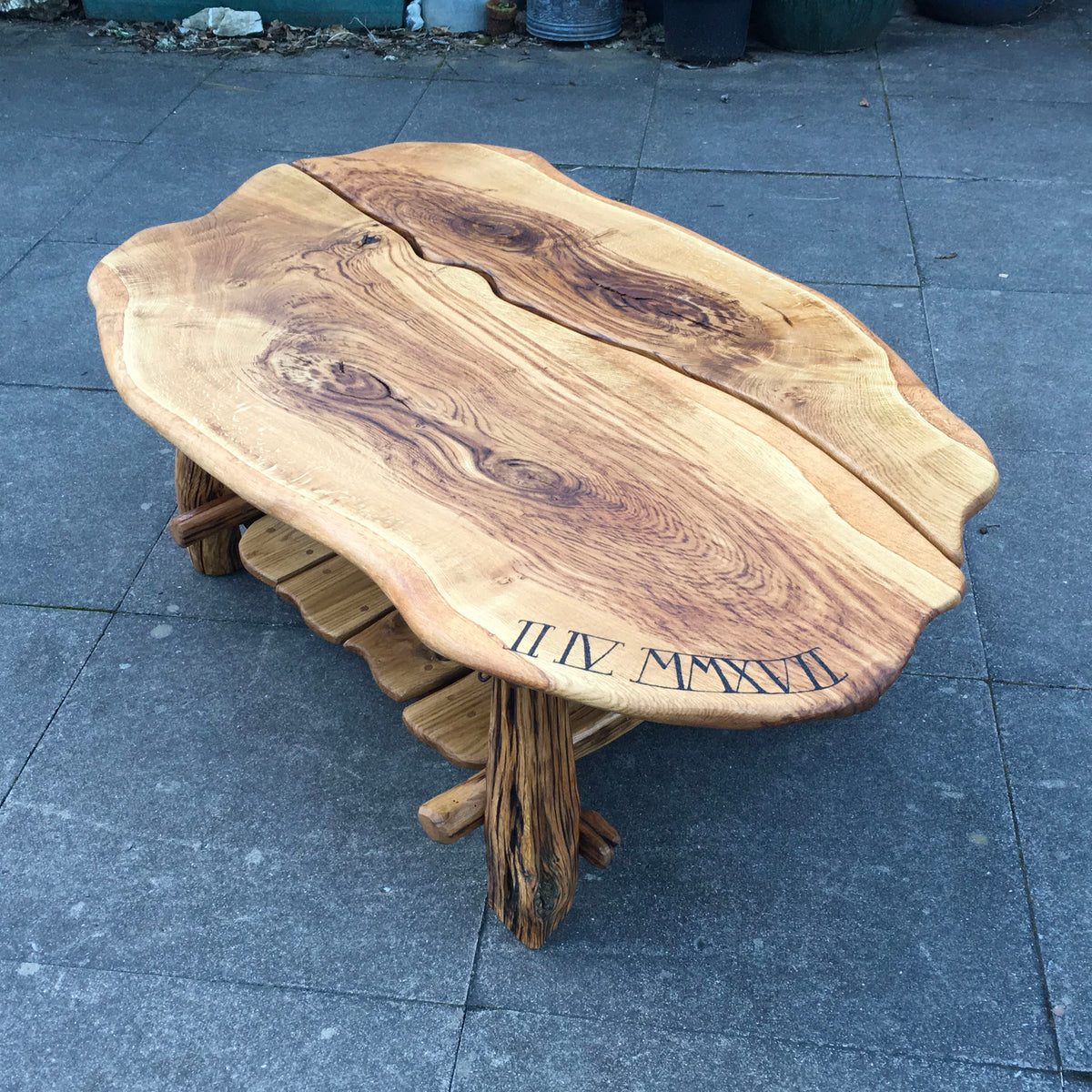 A rustic live-edge wooden coffee table with a natural bark-style base, photographed outdoors on a gray tiled floor. The tabletop has visible wood grain, a split center seam, and dark Roman numerals near one corner.